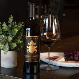 Bottle of Trinitas Cellars 2019 Cabernet Franc with wine glass, cheese, charcuterie, and grapes on a rustic table.
