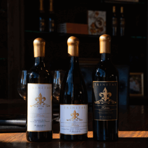 Fidelis Tasting – Now Available Three Trinitas Fidelis wine bottles displayed on a polished wooden table in warm ambient lighting.
