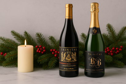 Two bottles of Trinitas wine, a Christmas Cuvée and a Brut Rosé, standing on a marble surface with pine branches and a glowing candle in a minimal holiday arrangement.