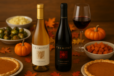 Two Trinitas wine bottles, Chardonnay and Pinot Noir, on a Thanksgiving table with fall leaves, pumpkin pie, mashed potatoes, and roasted vegetables.