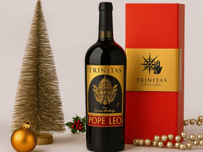 A bottle of Trinitas Cellars Pope Leo wine beside a red and gold Trinitas gift box, surrounded by festive holiday décor including gold ornaments, evergreen branches, and a shimmering Christmas tree on a neutral backdrop.