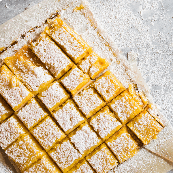 Lemon bars cut into squares, dusted with powdered sugar, on parchment paper with sugar scattered around.