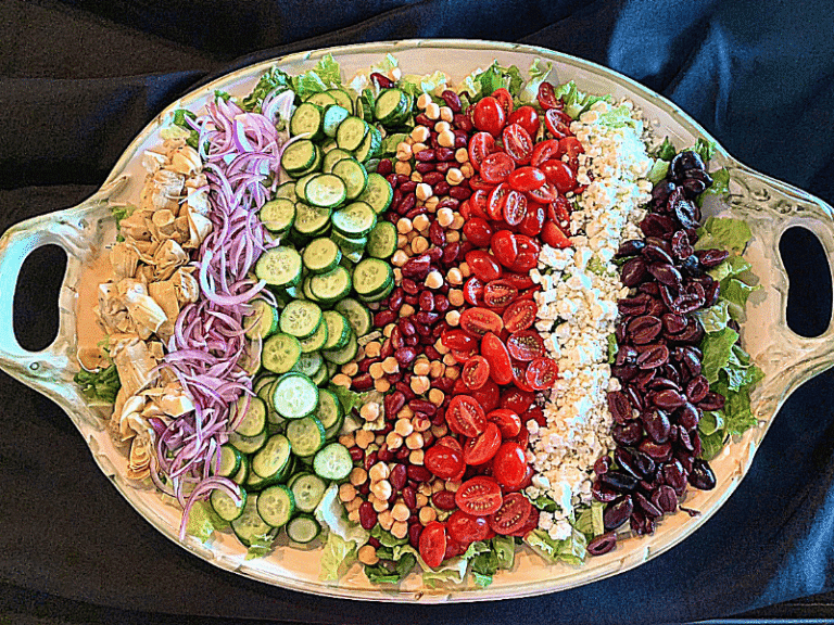 Large platter of colorful Greek salad with rows of veggies, feta cheese, and olives on a bed of lettuce.