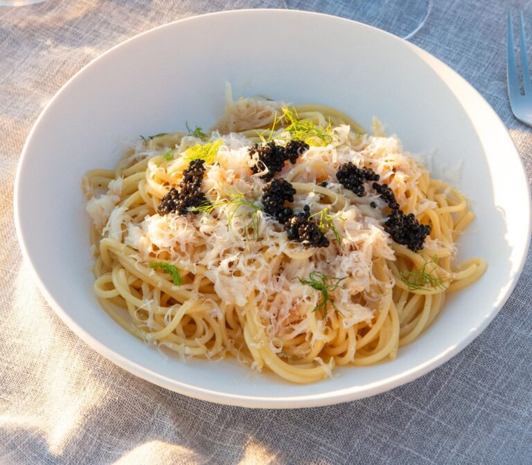 A bowl of spaghetti topped with grated cheese, herbs, and black caviar on a light tablecloth.
