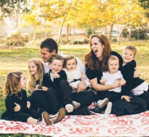 A family of seven sits on a picnic blanket outdoors, smiling and laughing together on a sunny day.