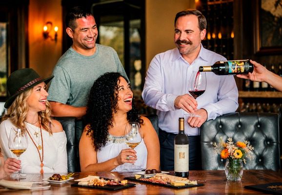 Four people at a table smiling and enjoying wine, with food platters and a bottle of wine in front of them.