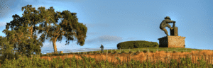 A large vineyard sculpture of a man opening a wine bottle stands on a hilltop beside a tree at sunset.