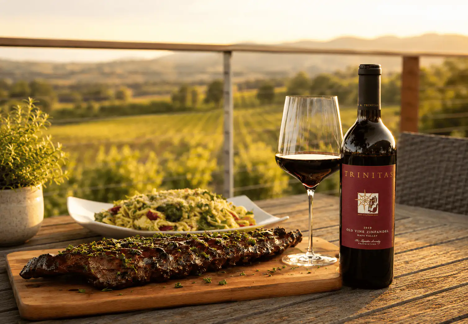 Grilled ribs, pasta salad, red wine, and glass on a table with a vineyard view at sunset.