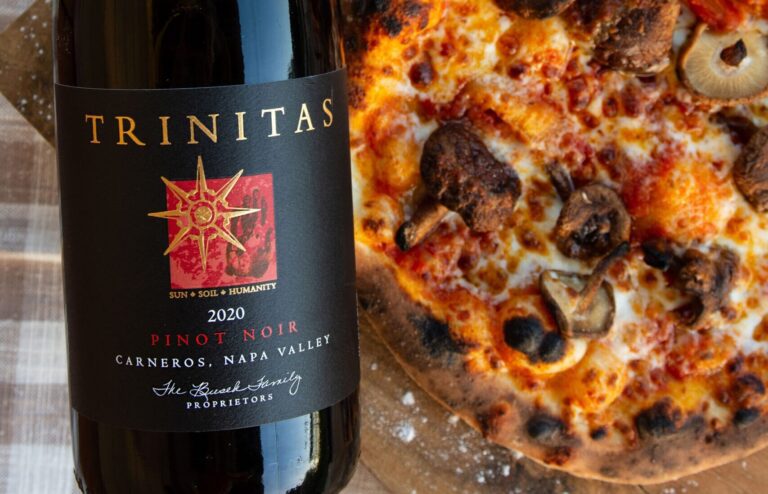 A bottle of Trinitas 2020 Pinot Noir next to a mushroom pizza on a wooden surface.