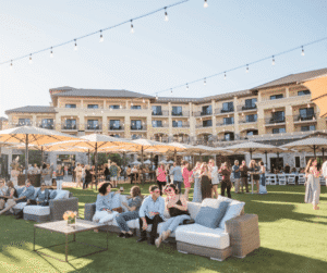 People gather and socialize outdoors on a lawn with couches, string lights, and a large hotel in the background.