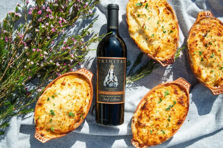 Bottle of red wine surrounded by four baked dishes and purple flowers on a light cloth background.
