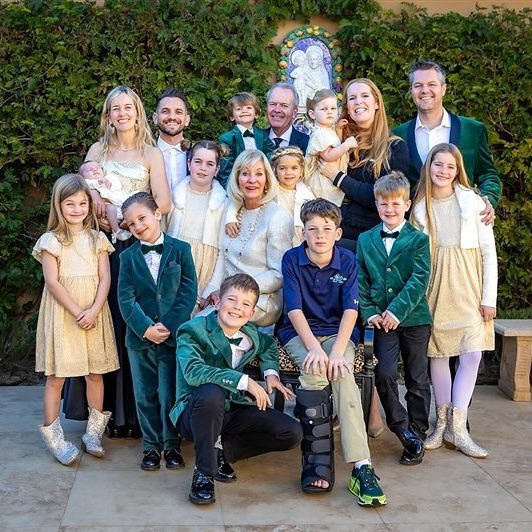 A large family poses outside in formal attire, smiling in front of green foliage and an angel decoration.