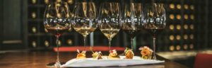 Five glasses of wine and an assortment of gourmet appetizers on a white plate, set on a wooden table.