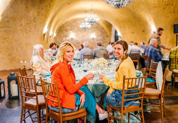Two women smiling and sitting at a table in a warmly lit, arched banquet hall with other guests dining.