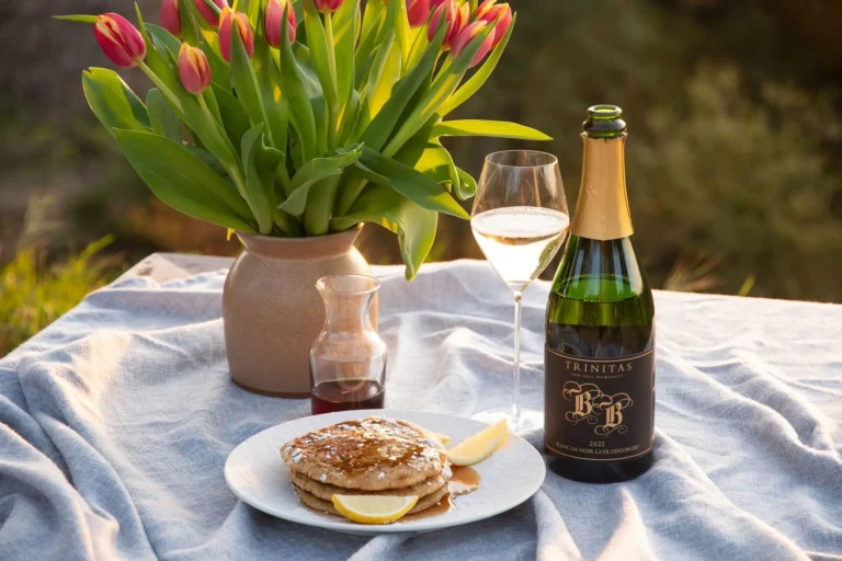 A plate of pancakes with lemon and syrup, a wine bottle, glass, and tulips on a picnic table.