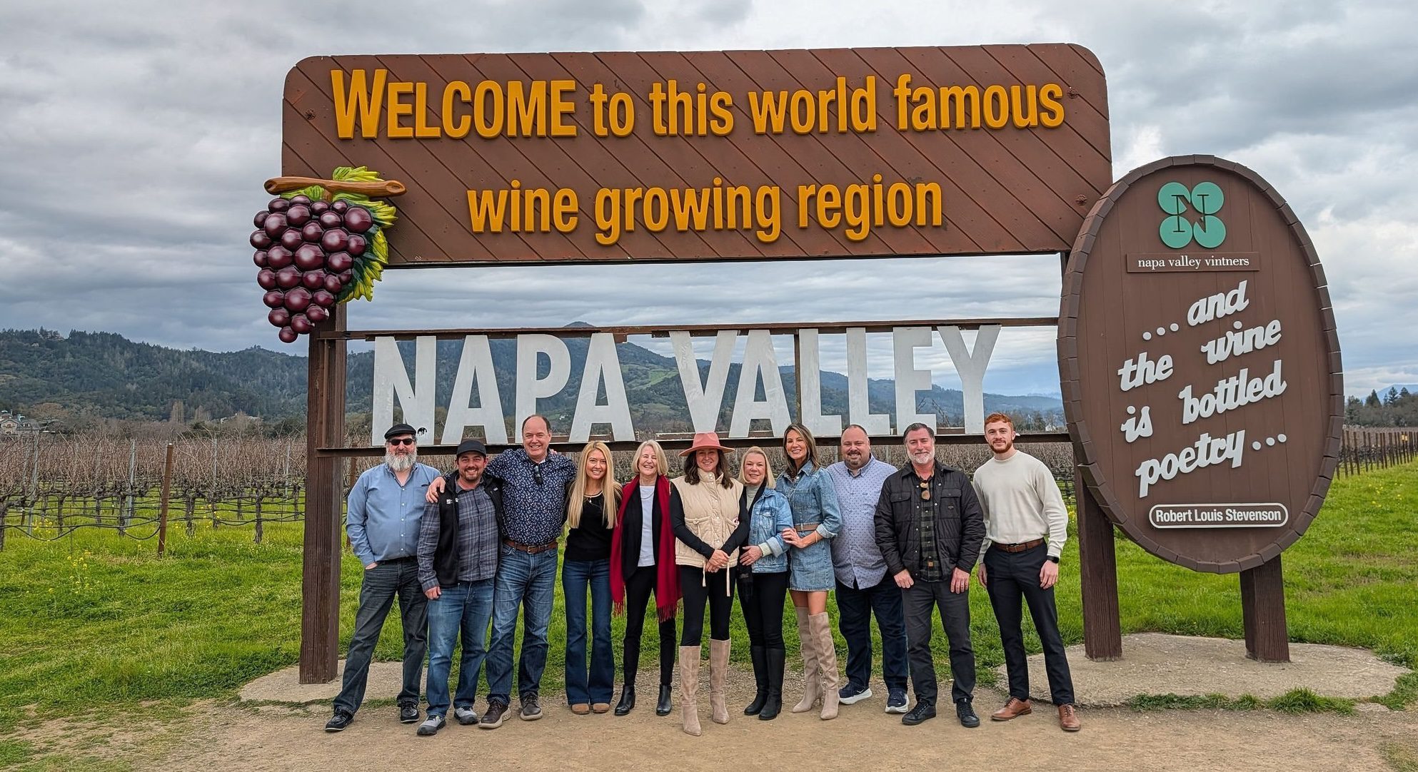Ten people pose smiling in front of a large Welcome to Napa Valley wine region sign with vineyards and hills behind.