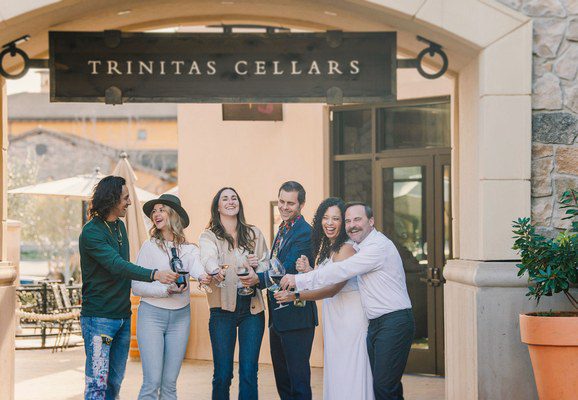 Six people smiling with wine glasses under a Trinitas Cellars sign at a winery entrance.