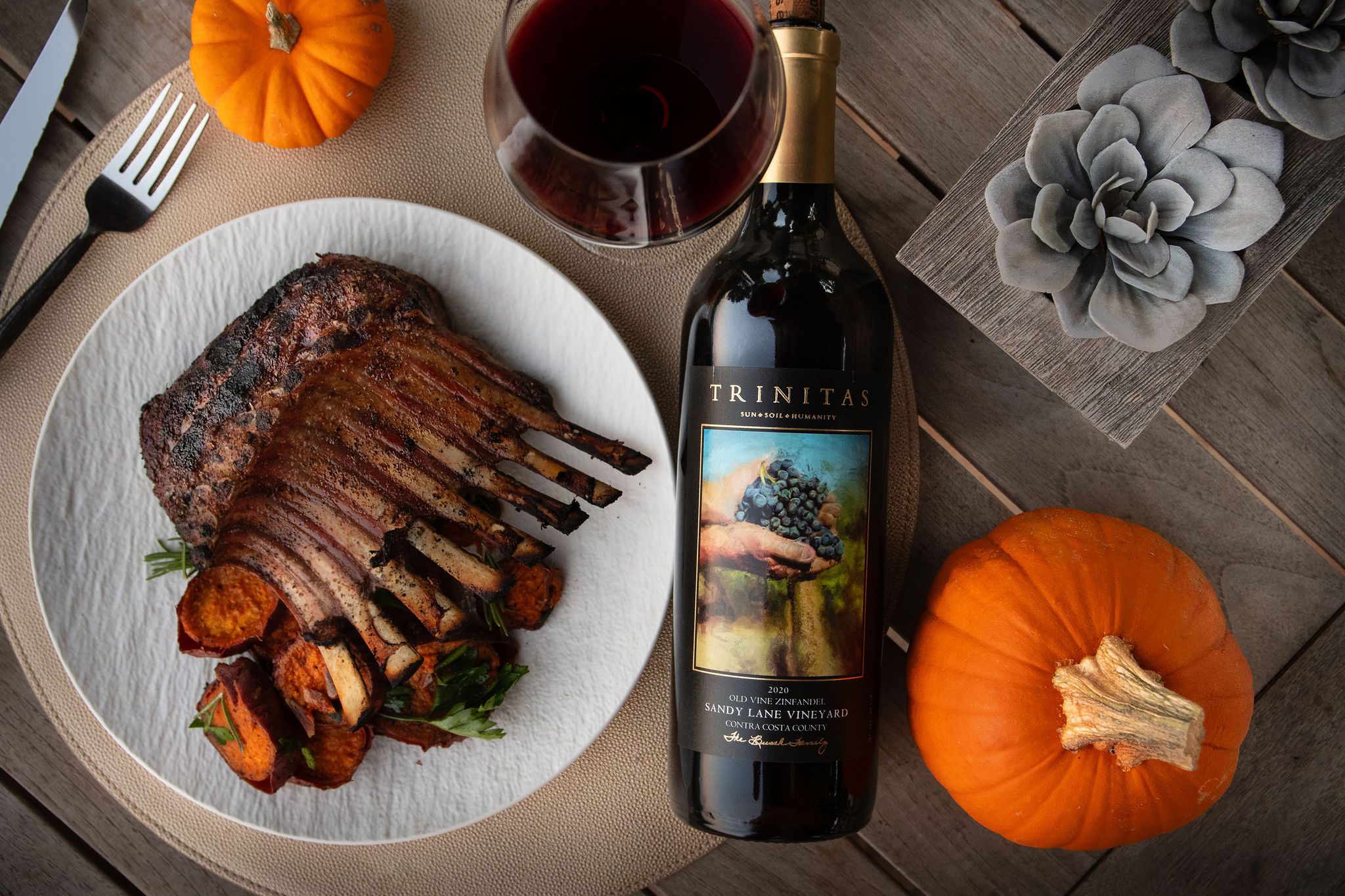 Sandy Lane Old Vine Zinfandel bottle beside grilled ribs, a glass of red wine, pumpkins, and seasonal table details.