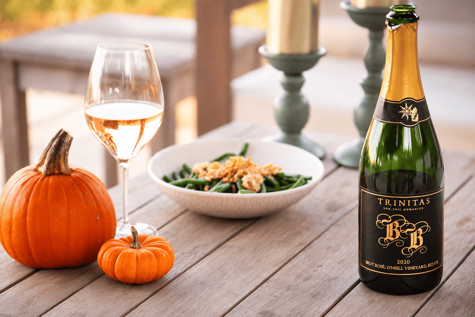 A bottle of Napa wine, a glass, two pumpkins, and a bowl of green beans rest on a wooden table.