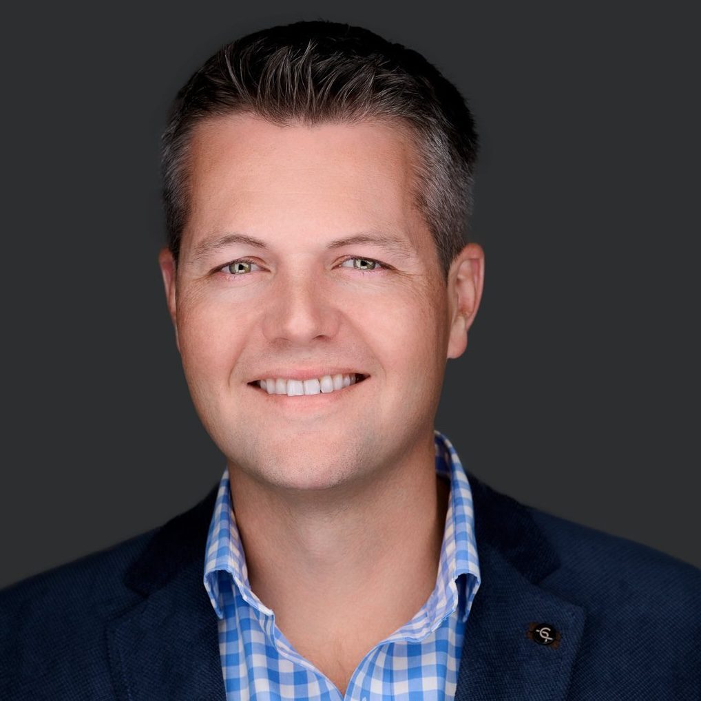 Smiling man in a blue checked shirt and dark blazer against a plain dark background.