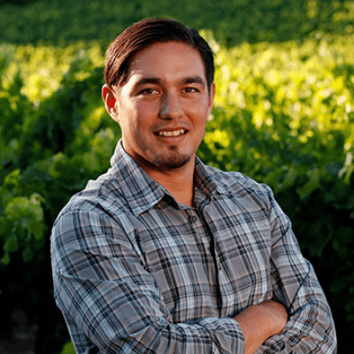 Man in a plaid shirt standing with arms crossed in a vineyard, smiling at the camera.