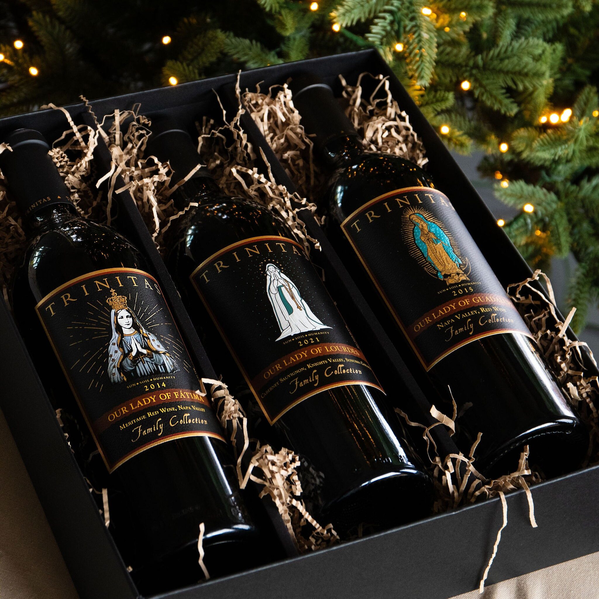 Three Trinitas Cellars Marian Series wines — Our Lady of Guadalupe, Our Lady of Lourdes, and Our Lady of Fatima — elegantly boxed under a Christmas tree with twinkling lights.