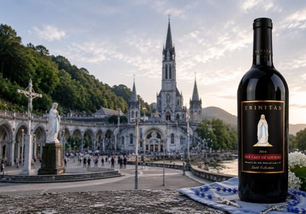 A bottle of Trinitas Christian Wine in front of Lourdes Sanctuary in France at sunset, surrounded by people and vibrant flowers.