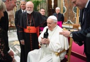 The Pope sits smiling, holding a wine bottle, surrounded by clergy and others in a grand room.