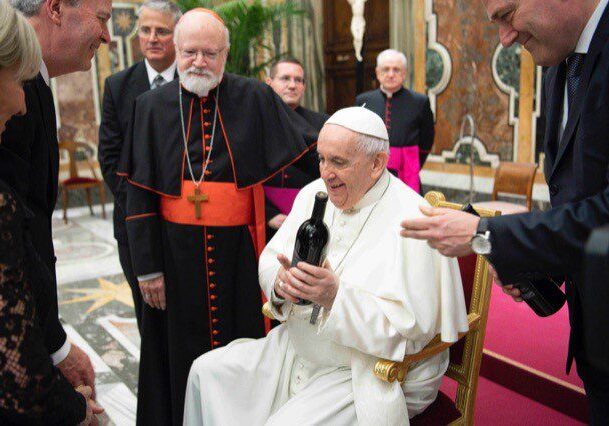 The Pope sits smiling, holding a wine bottle, surrounded by clergy and others in a grand room.