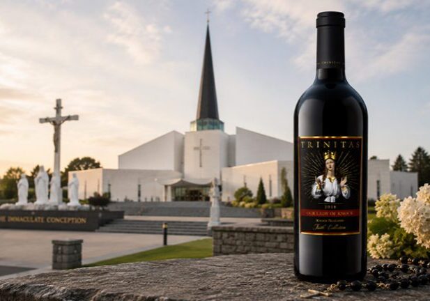 A bottle of Christian Wine stands on a stone ledge before a church, with a rosary and flowers beside it at sunset.