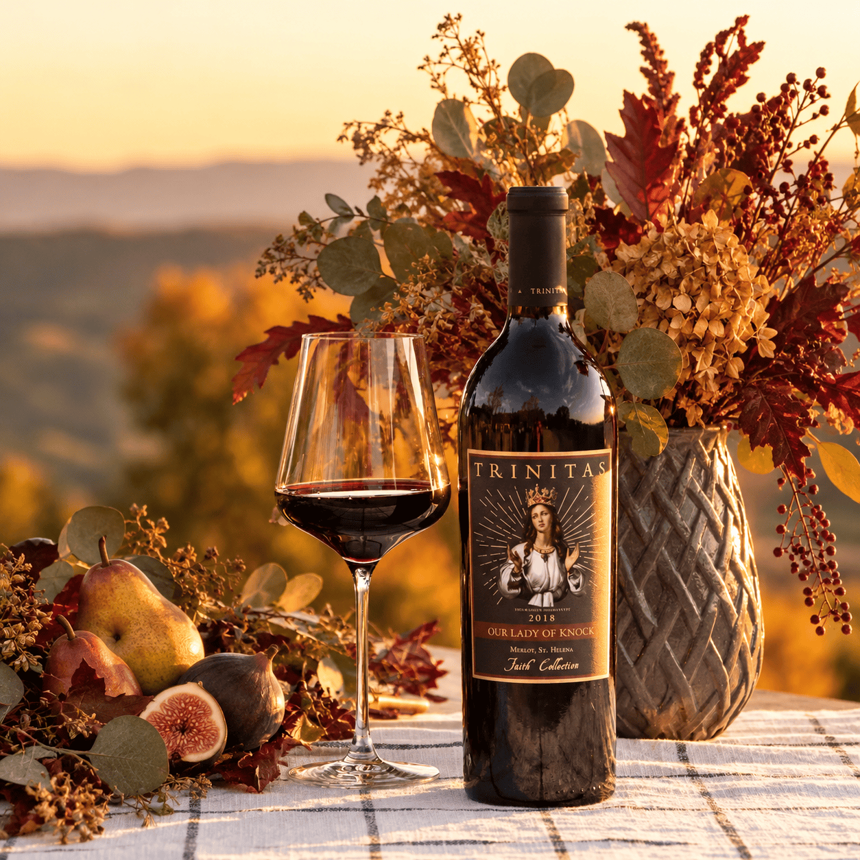 Trinitas Our Lady of Knock Merlot bottle and red wine glass surrounded by autumn foliage, figs, and pears.