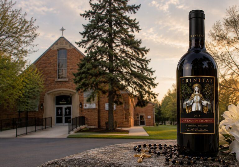 A Trinitas Christian Wine bottle rests on a stone, rosary nearby, with a church building in the background at sunset.
