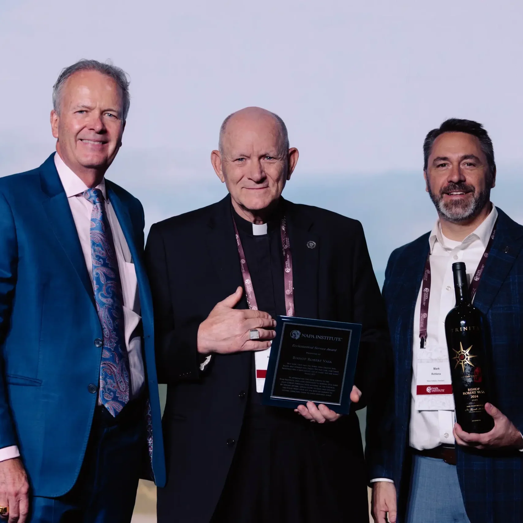 Three men in suits pose together; one holds a plaque and another holds a large wine bottle.