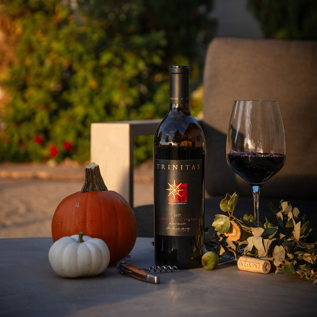 Trinitas Cabernet Sauvignon bottle and glass with pumpkins and fall decor on a patio table