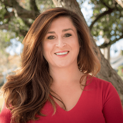 Woman with long brown hair in a red top smiling outdoors with trees in the background.