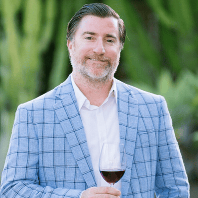 Man in a light blue plaid blazer holds a glass of red wine, standing outdoors with greenery in the background.