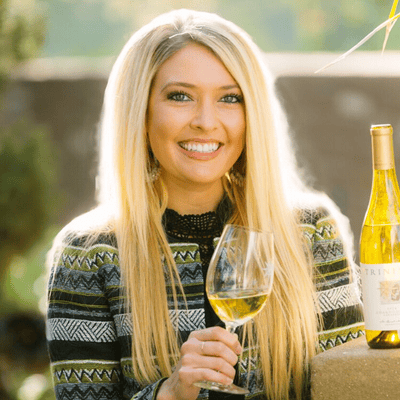 Smiling woman with long blonde hair holding a glass of white wine next to a wine bottle outdoors.