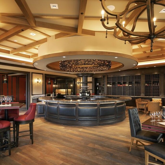 Elegant restaurant interior with round central bar, wine rack, wooden beams, and red and black upholstered chairs.