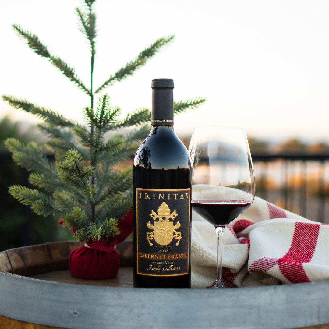 A bottle of Trinitas red wine beside a small decorated Christmas tree and a glass of wine on a barrel table, wrapped in a cozy red-and-white blanket at sunset.