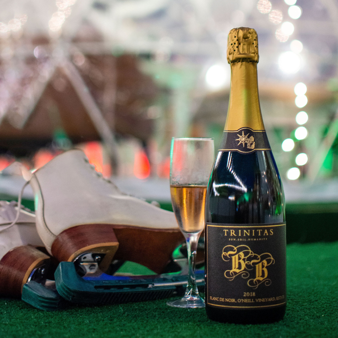 A bottle of Trinitas Brut Sparkling with a filled champagne flute beside a pair of ice skates, set against a softly lit winter backdrop.