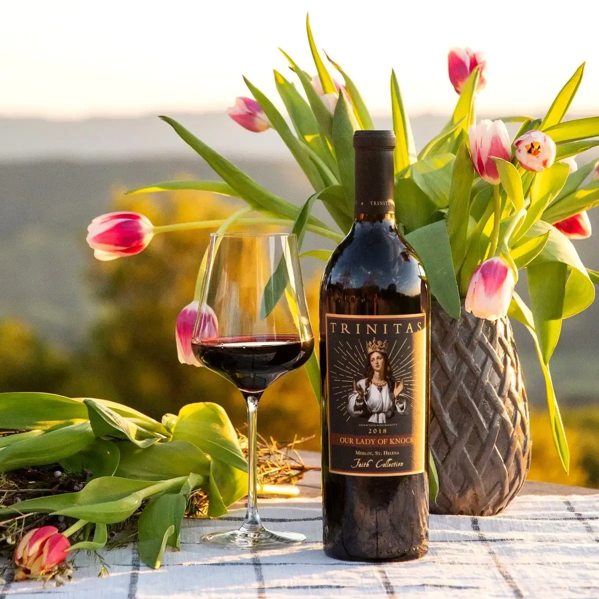 A bottle of Trinitas wine and a glass of red wine beside a vase of tulips on an outdoor table at sunset.
