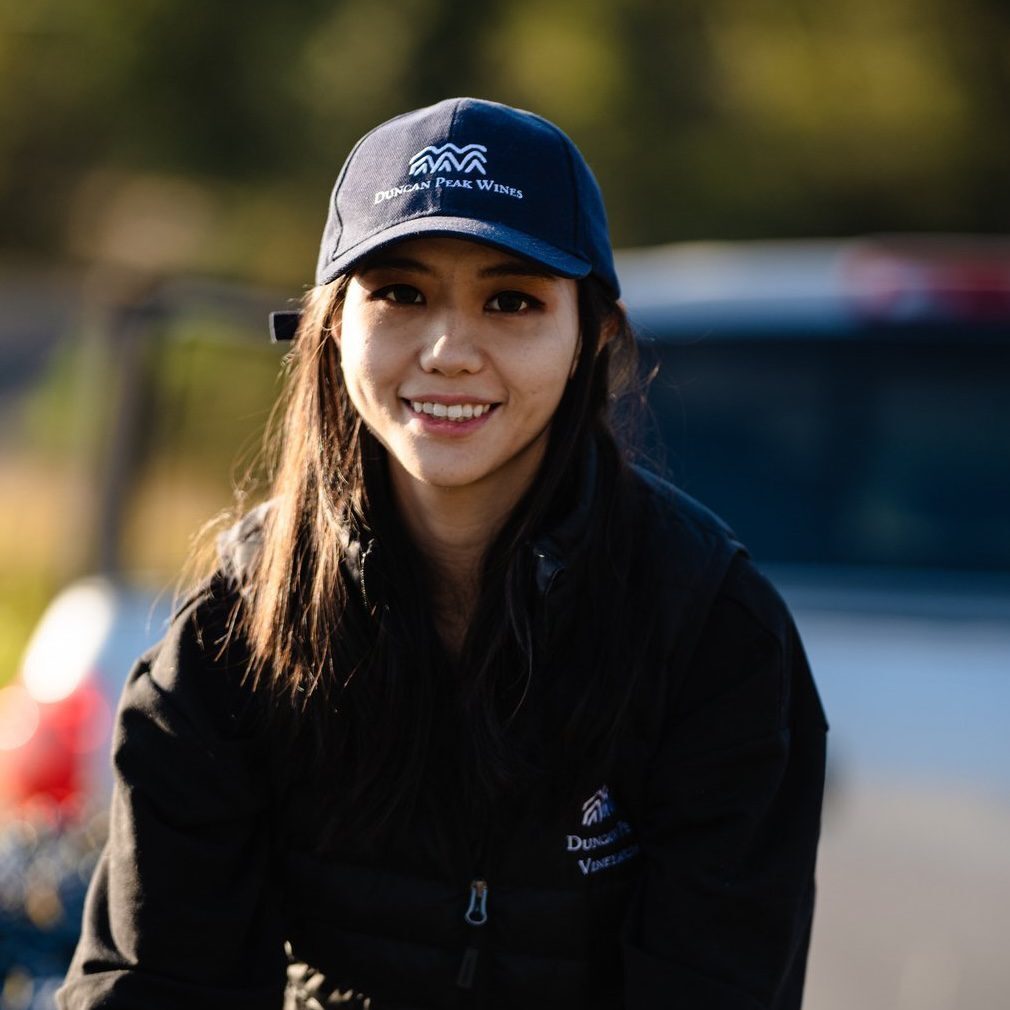 A woman in a logo hat and vest smiles outdoors with a blurred car in the background.