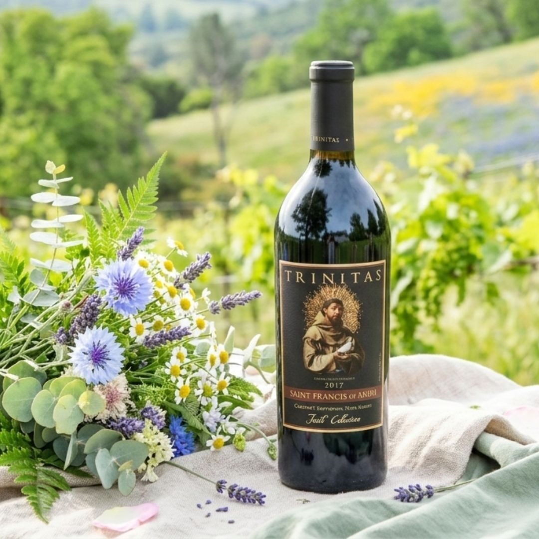 A bottle of Trinitas red wine next to a bouquet of wildflowers outdoors on a picnic blanket.
