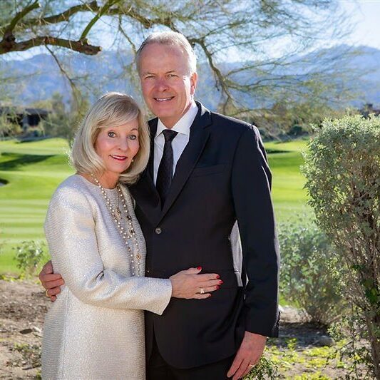 Smiling older couple in formal attire posing outdoors on a sunny day with greenery and mountains behind them.